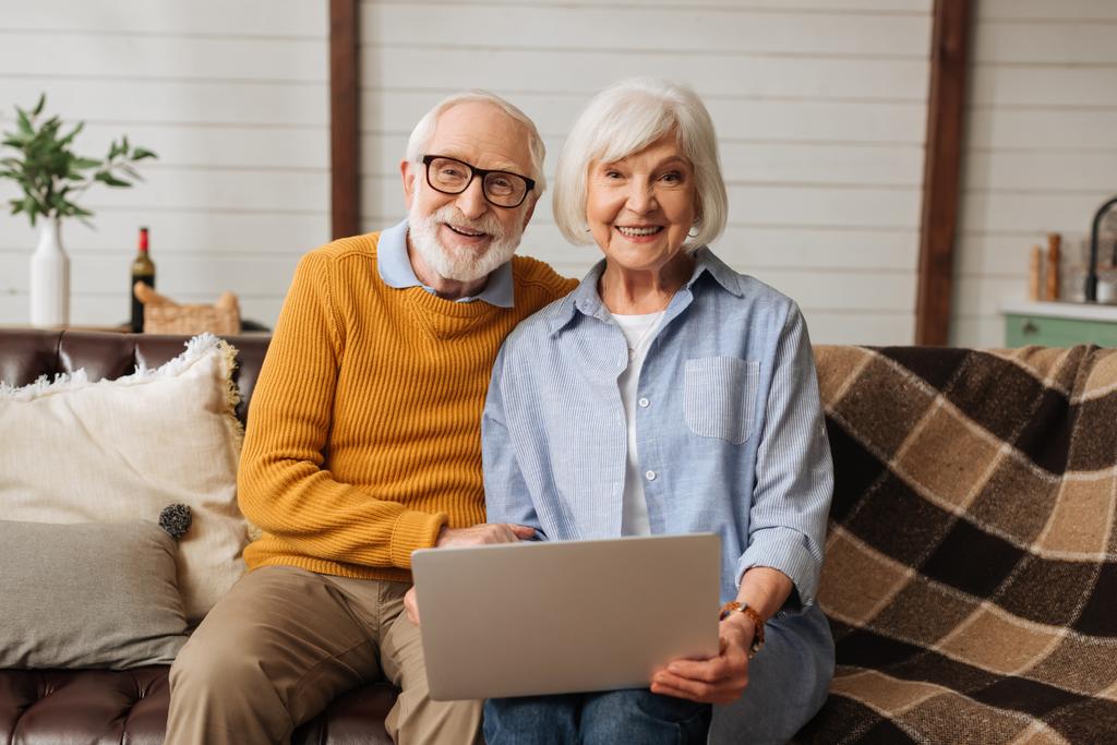 cheerful elderly husband hugging wife with laptop while looking at camera on couch in living room on blurred background - Photo, Image