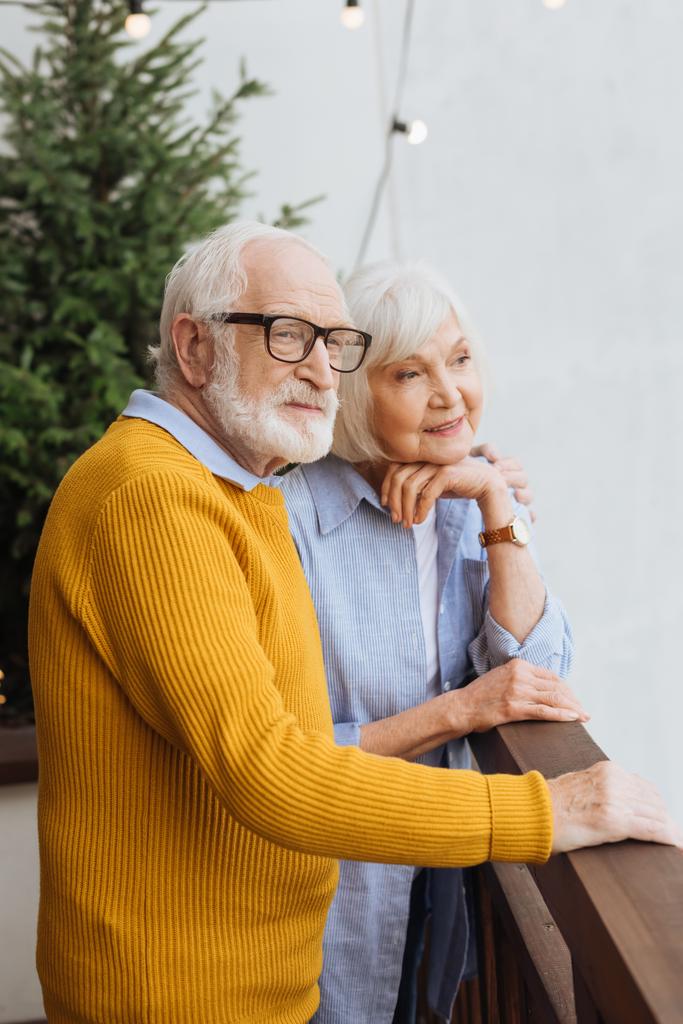 idős férj ölelés mosolygós feleség, miközben néz el a teraszon homályos háttér - Fotó, kép