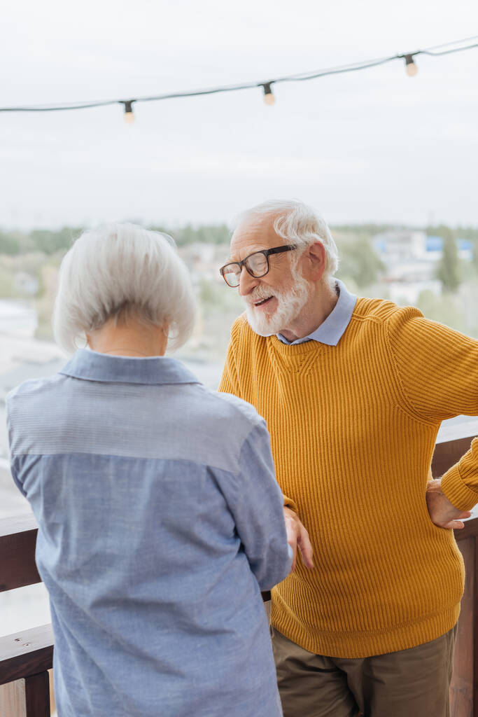 visão traseira da esposa idosa de pé perto do marido sorridente com a mão no quadril no terraço no fundo borrado - Foto, Imagem