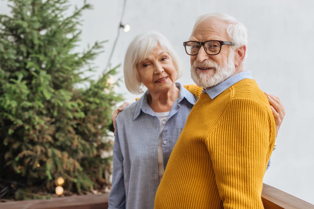 glückliches älteres Paar blickt in die Kamera, während es sich auf der Terrasse vor verschwommenem Hintergrund umarmt - Foto, Bild