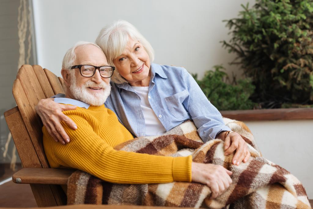 feliz pareja de ancianos con manta a cuadros mirando a la cámara mientras se abraza en el sillón sobre fondo borroso en la terraza - Foto, Imagen