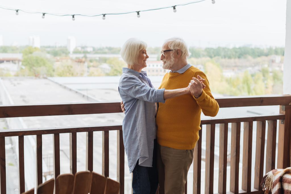 šťastný starší pár při pohledu na sebe při tanci na terase na rozmazaném pozadí - Fotografie, Obrázek