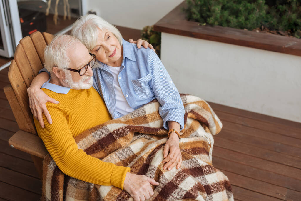 улыбающаяся пожилая пара с пледом одеяло, обнимая и глядя в сторону, сидя в кресле на террасе на размытом фоне - Фото, изображение