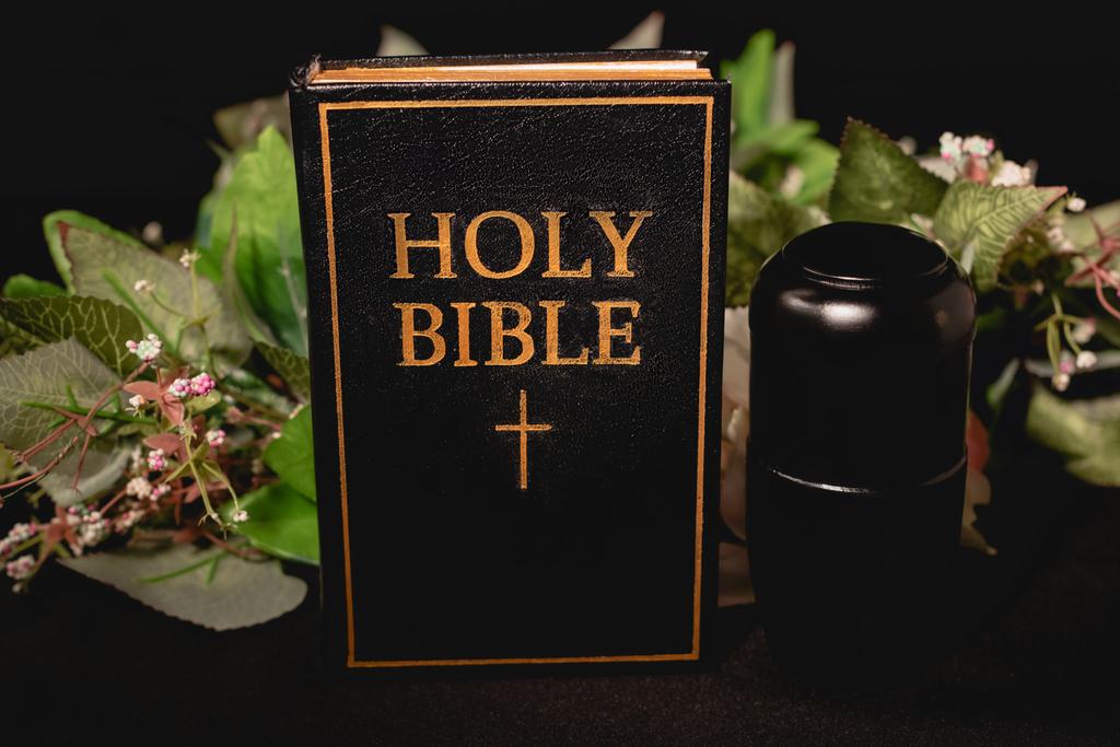 Святая Библия и урна с пеплом на черном фоне, похоронная концепция - Фото, изображение