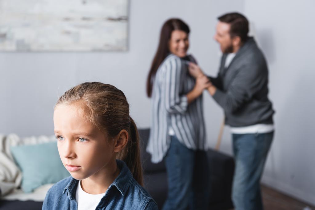 Αναστατωμένο παιδί που στέκεται κοντά σε γονείς που μαλώνουν σε θολή φόντο στο σπίτι  - Φωτογραφία, εικόνα