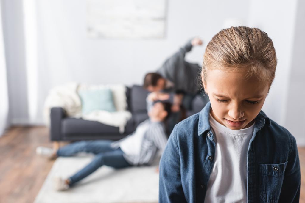 Weinendes Mädchen steht neben Vater und schlägt Mutter auf dem Boden im Wohnzimmer auf verschwommenem Hintergrund  - Foto, Bild