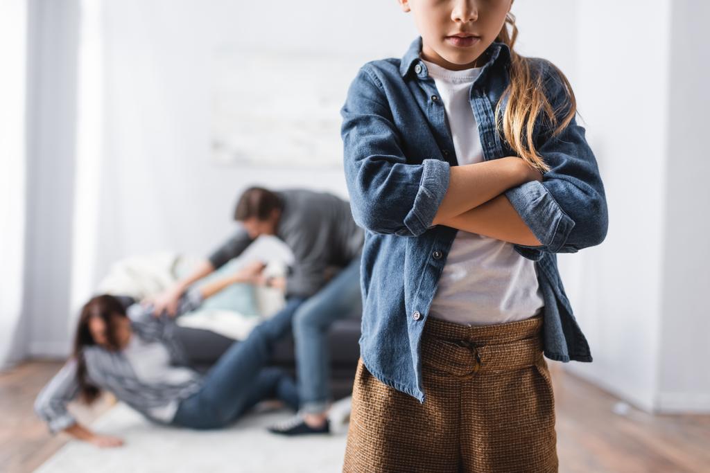 Chica con brazos cruzados de pie cerca de padre abusivo golpeando a la madre en casa en un fondo borroso  - Foto, Imagen