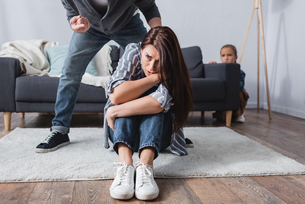 Donna spaventata seduta sul pavimento vicino al marito abusivo che punta con il dito e la figlia nascosti dietro il divano su sfondo sfocato  - Foto, immagini