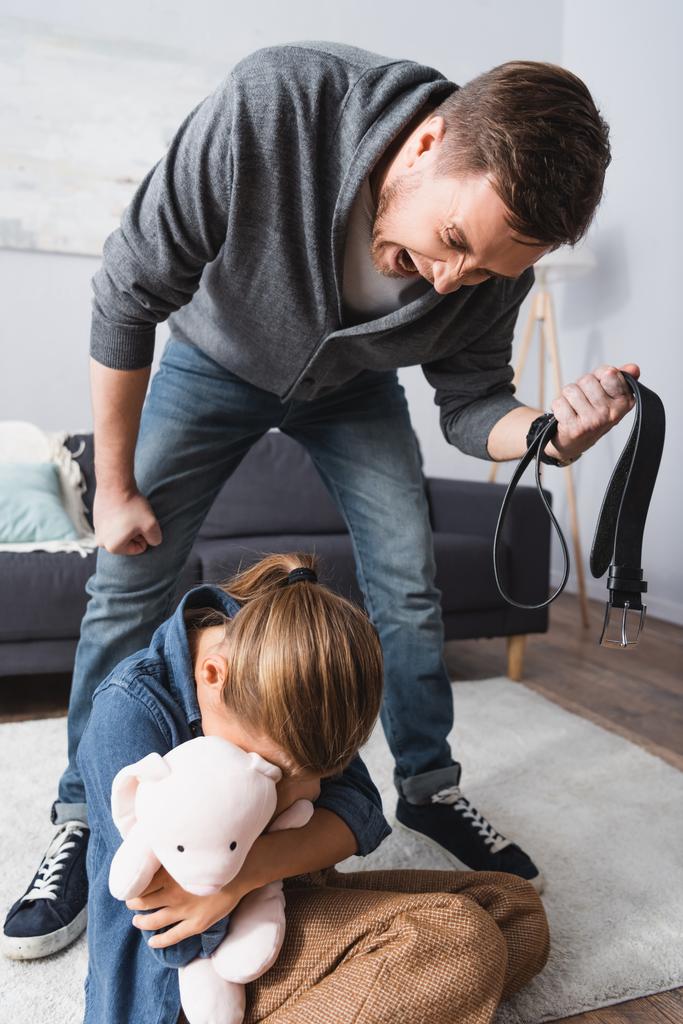 Злой мужчина с поясом кричит на дочь с мягкой игрушкой дома  - Фото, изображение