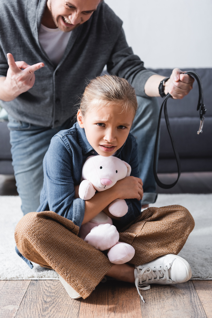 Przestraszone dziecko trzymające miękką zabawkę w pobliżu wściekłego ojca z paskiem w pasie na rozmytym tle  - Zdjęcie, obraz