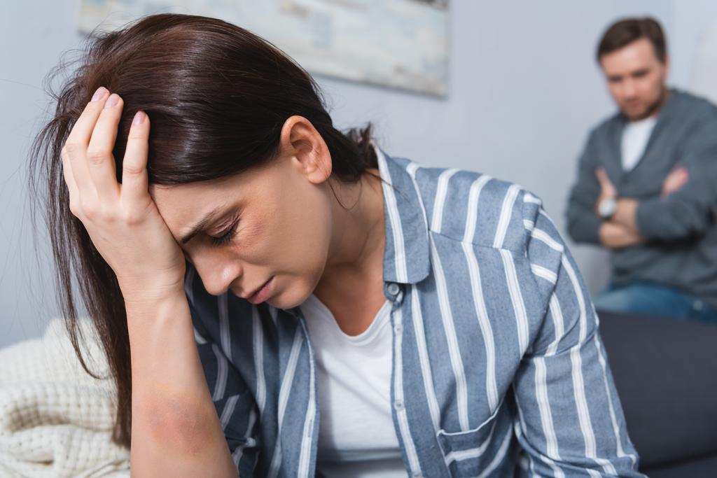 depresivní žena s modřinou v blízkosti oka sedí v blízkosti zneužívající manžel na rozmazaném pozadí doma  - Fotografie, Obrázek