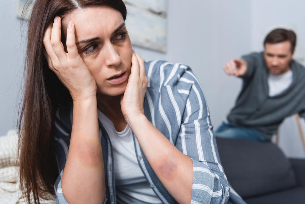 beledigde vrouw met blauwe plekken zitten in de buurt van misbruikende echtgenoot wijzend met de vinger op wazig achtergrond  - Foto, afbeelding