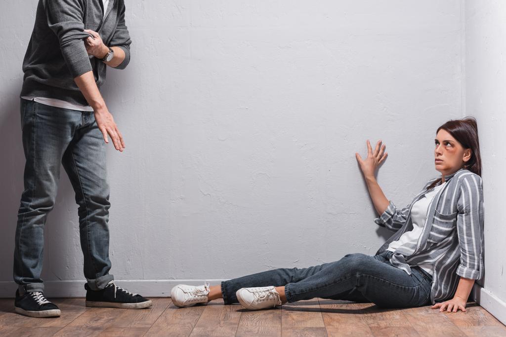 Bang vrouw met blauwe plekken zittend op de vloer in de buurt van misbruiker oprollen mouwen  - Foto, afbeelding