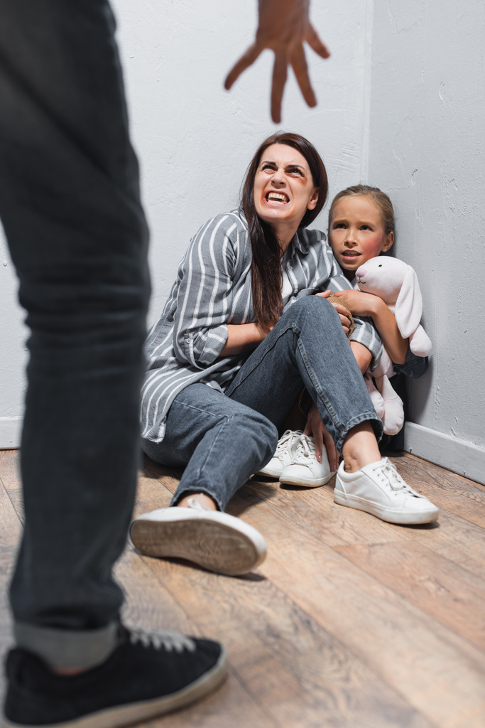 Screaming woman with bruises and kid with soft toy looking at abusive father on blurred foreground  - Photo, Image