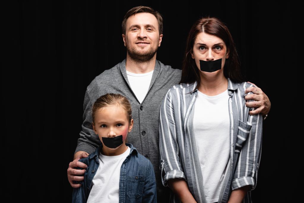 Smiling abuser hugging wife and daughter with bruises and adhesive tapes isolated on black   - Photo, Image