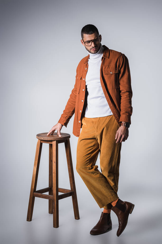 Модный мужчина, стоящий возле деревянного стула на сером фоне  - Фото, изображение