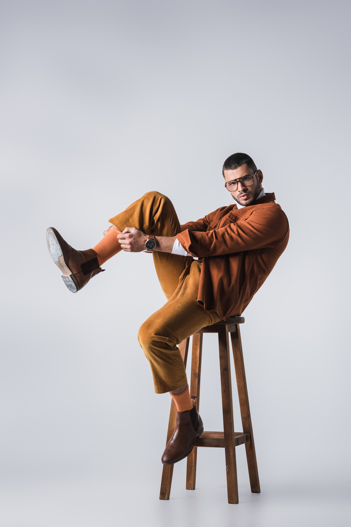 Modieuze man die oranje sok aanpast terwijl hij op een stoel op een grijze achtergrond zit  - Foto, afbeelding
