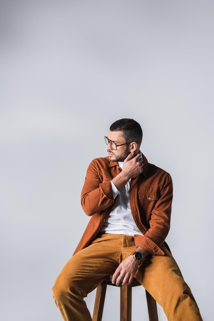 Mladý muž v neformálních šatech a brýlích odvrací pohled na židli izolované na šedé  - Fotografie, Obrázek