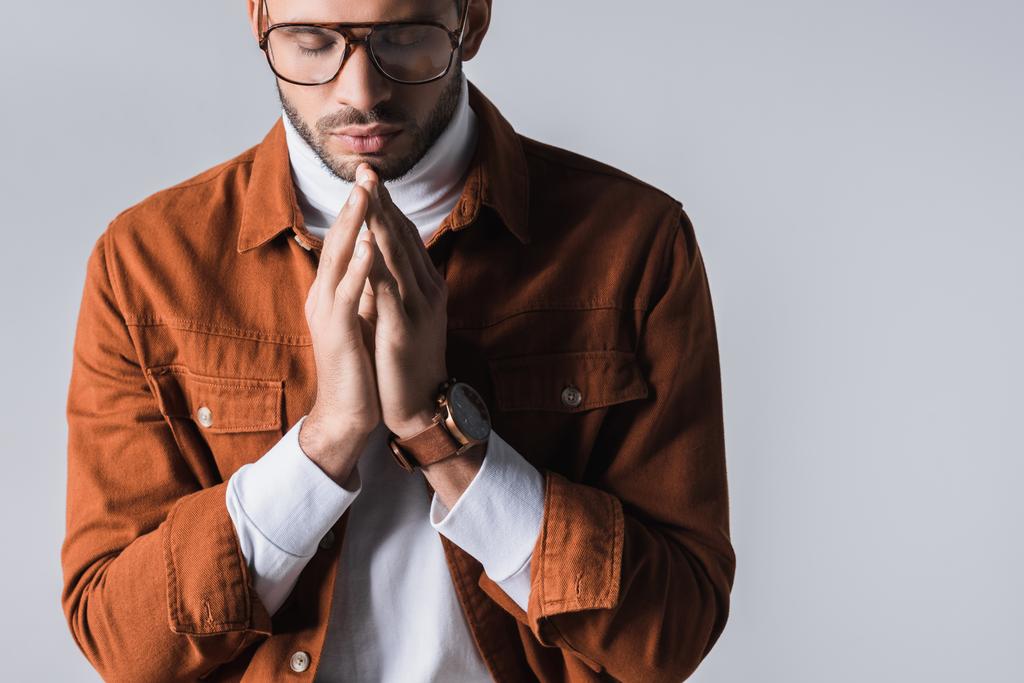 Tyylikäs mies, jolla on suljetut silmät, osoittaa rukoilevia käsiä harmaalla  - Valokuva, kuva