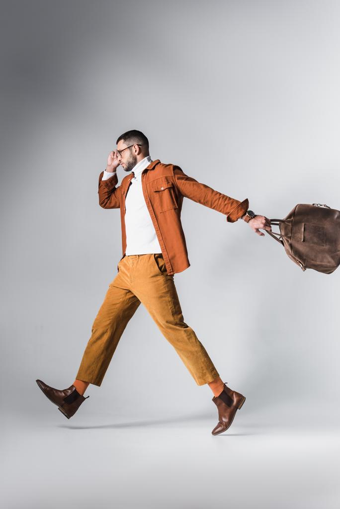 Hombre de moda sosteniendo bolsa marrón mientras camina sobre fondo gris  - Foto, Imagen