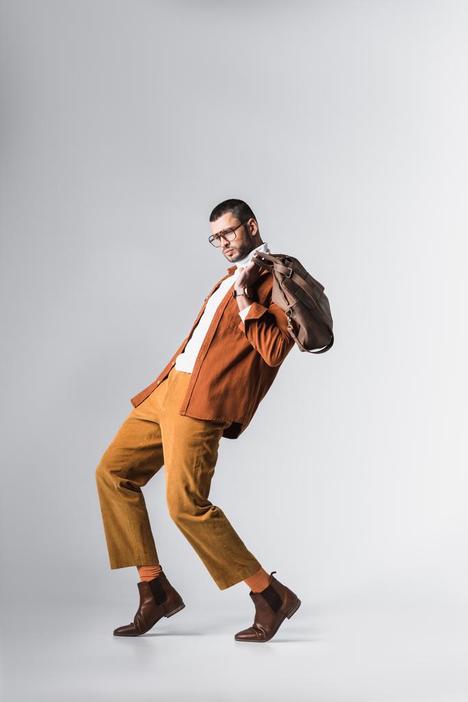 Man in terracotta jacket and brown παντελόνι κρατώντας τσάντα σε γκρι φόντο  - Φωτογραφία, εικόνα