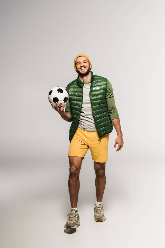 Χαμογελώντας αθλητής κρατώντας το ποδόσφαιρο, ενώ το περπάτημα σε γκρι φόντο  - Φωτογραφία, εικόνα