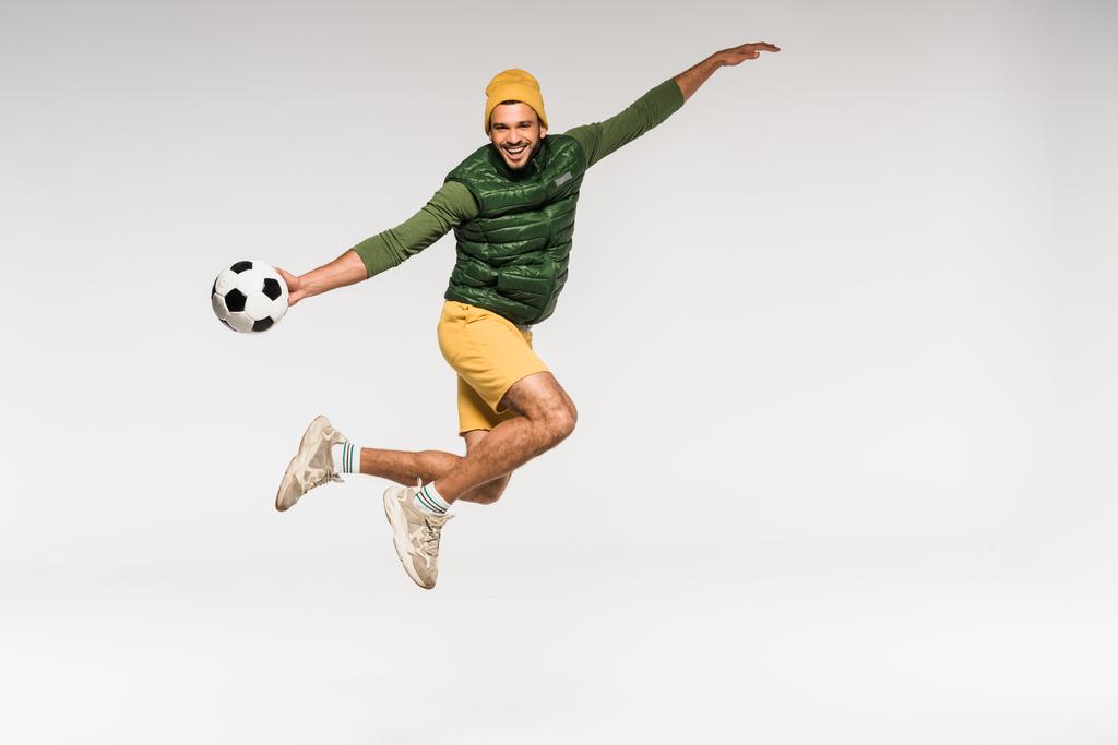 Allegro sportivo in abiti casual che salta e tiene il calcio isolato sul grigio  - Foto, immagini