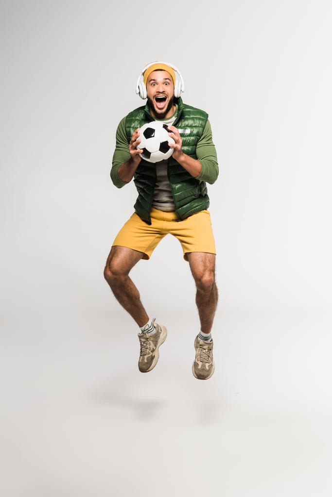 Захоплений спортсмен в навушниках стрибає і тримає футбол на сірому фоні
  - Фото, зображення