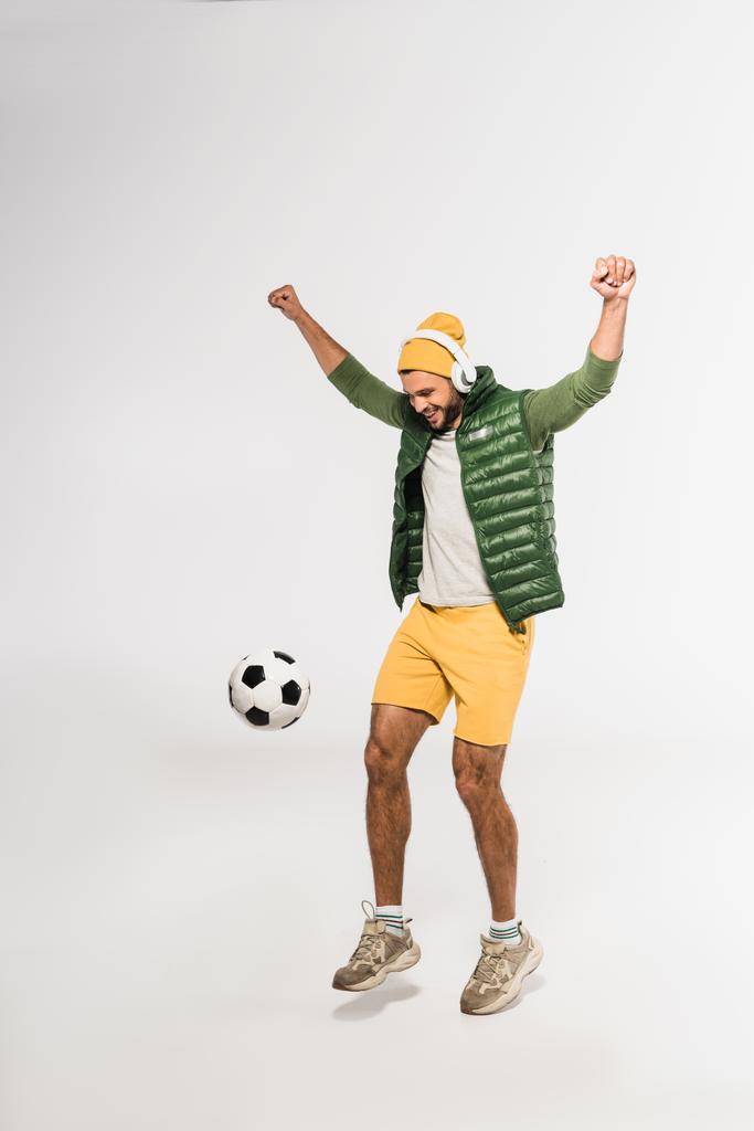 Opgewonden sportman in hoofdtelefoon tonen ja gebaar tijdens het spelen van voetbal op witte achtergrond - Foto, afbeelding