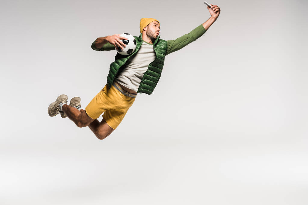 Sportowiec z piłką nożną robi selfie na smartfonie skacząc na szarym tle  - Zdjęcie, obraz