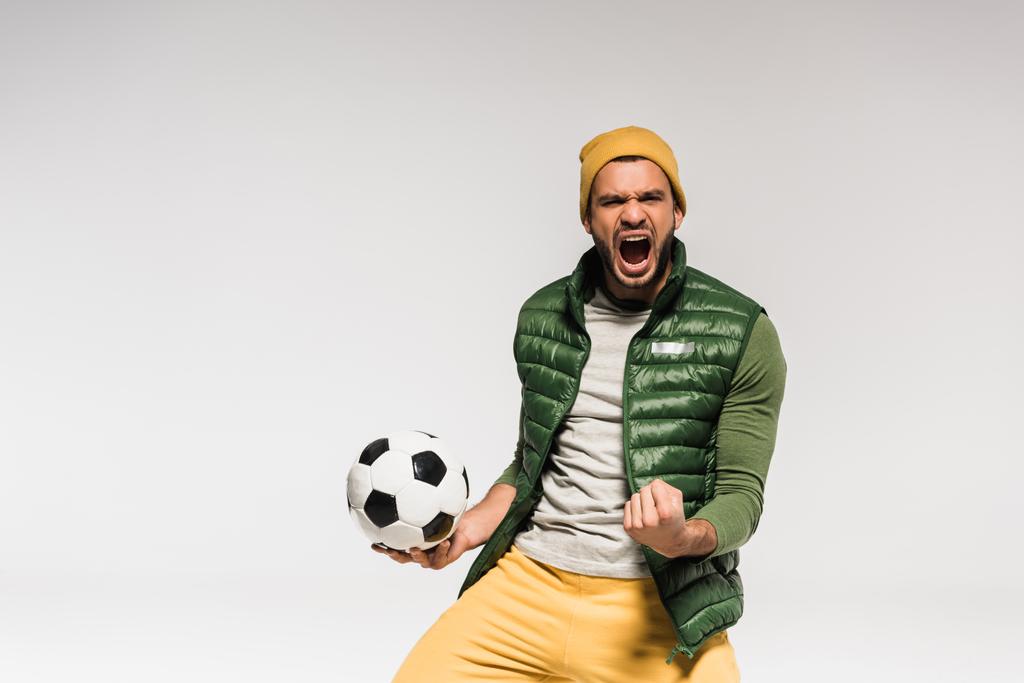 Ενθουσιασμένος αθλητής με ανοιχτό στόμα που δείχνει ναι χειρονομία και κρατώντας το ποδόσφαιρο απομονωμένο σε γκρι  - Φωτογραφία, εικόνα