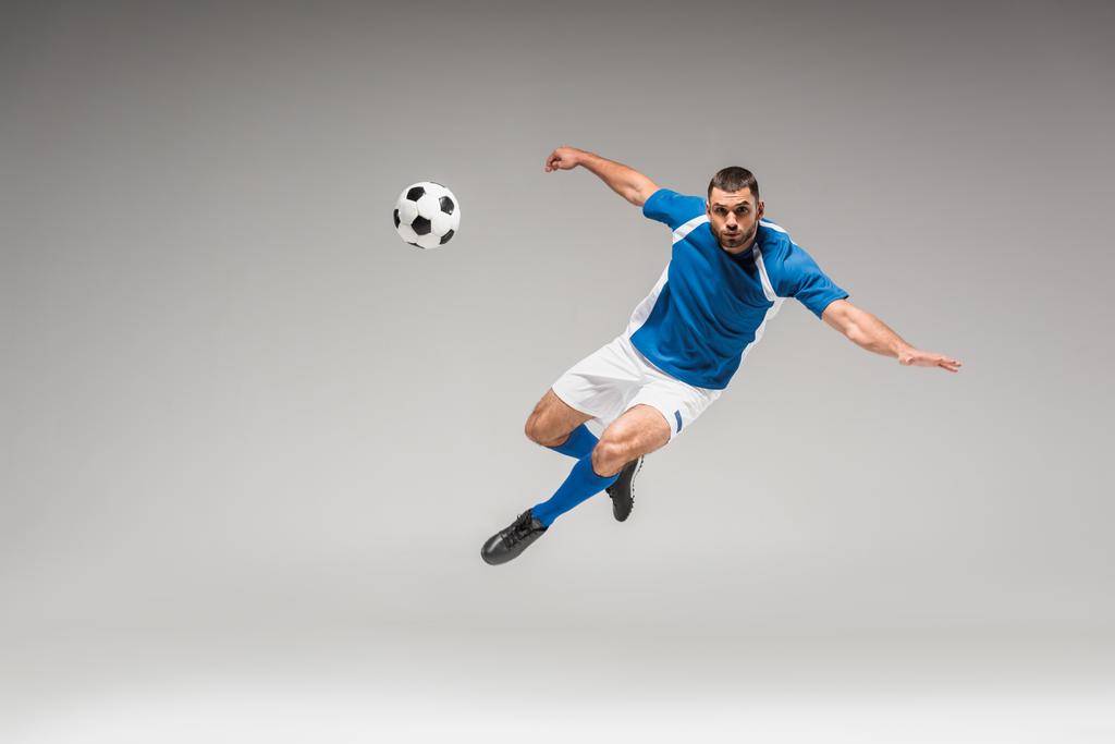 Sportowiec patrząc w kamerę podczas skoków w pobliżu piłki nożnej na szarym tle - Zdjęcie, obraz