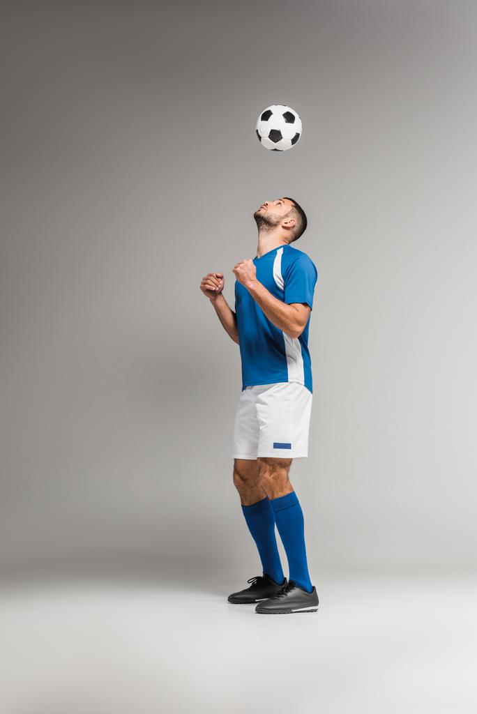 Молодой спортсмен смотрит на футбол в воздухе на сером фоне  - Фото, изображение