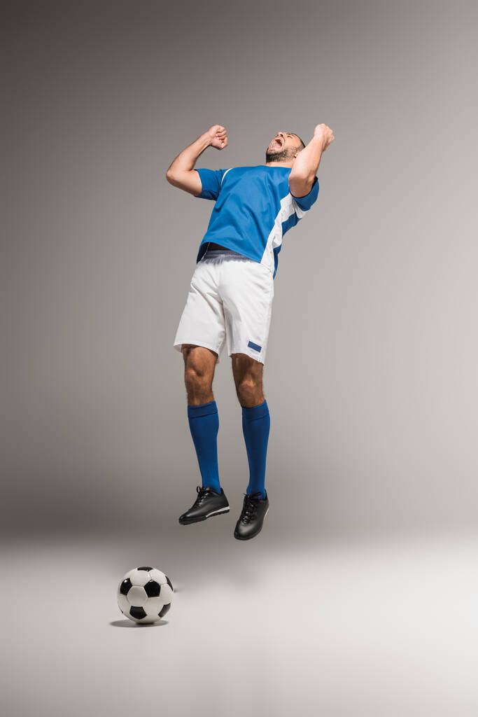 Amazed sportsman showing yes gesture while jumping near football on grey background  - Photo, Image