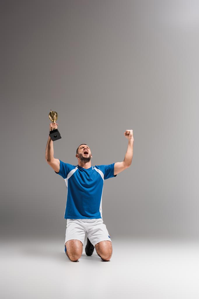 Deportista con copa campeona dorada mostrando sí gesto mientras se arrodilla sobre fondo gris  - Foto, Imagen