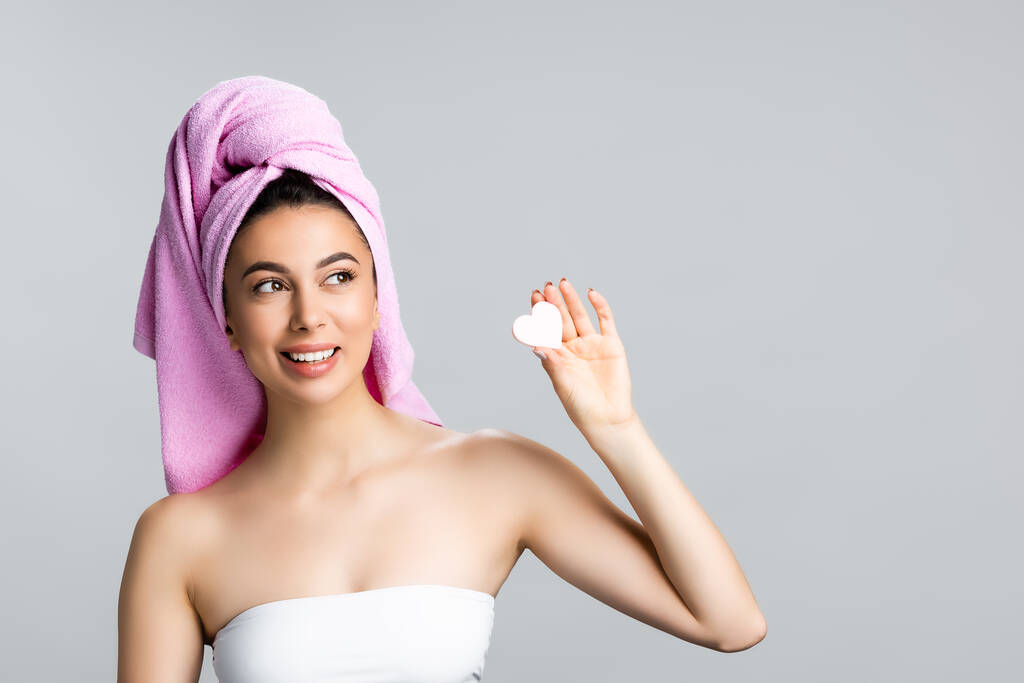 happy beautiful woman with towel on hair holding heart shaped sponge isolated on grey - Photo, Image
