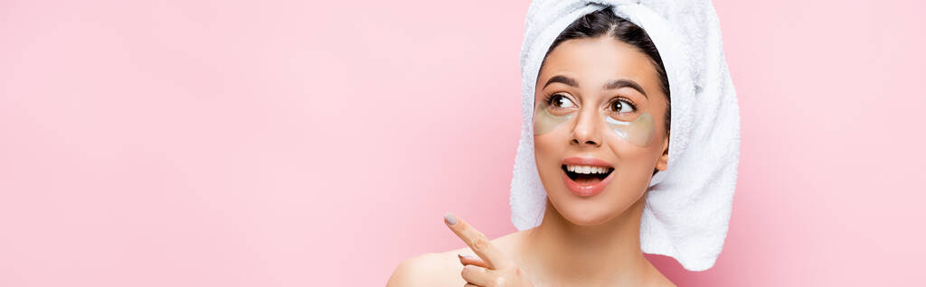 opgewonden mooie vrouw met handdoek op haar en hydrogel oogvlekken op gezicht geïsoleerd op roze, banner - Foto, afbeelding