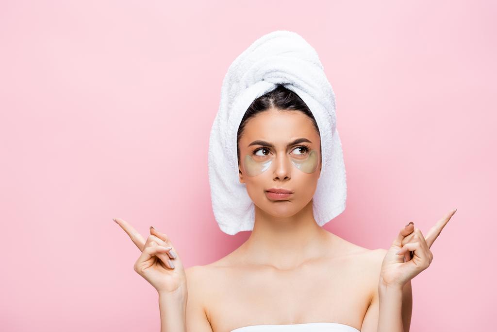 hermosa mujer pensativa con toalla en el cabello y parches de hidrogel en la cara señalando con los dedos aislados en rosa - Foto, Imagen
