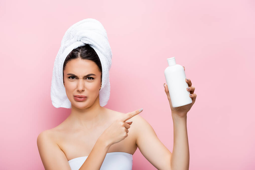 disgusted beautiful woman with towel on hair pointing at bottle of lotion isolated on pink - Photo, Image