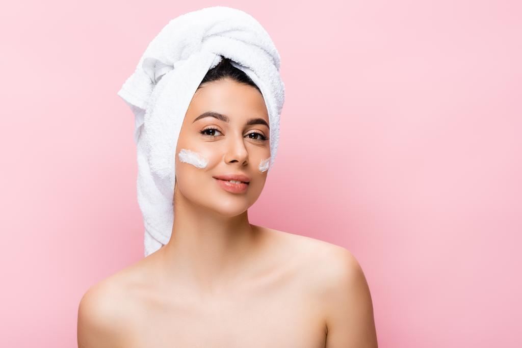 mooie vrouw met handdoek op haar en cosmetische crème op gezicht geïsoleerd op roze - Foto, afbeelding