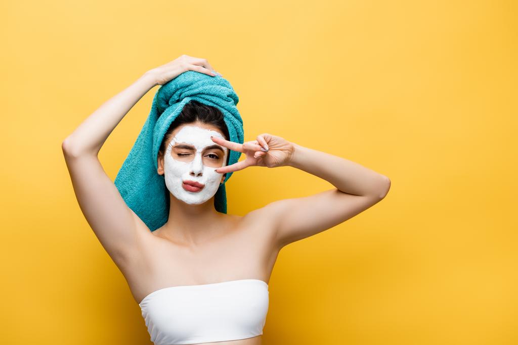 beautiful woman with blue towel on hair and clay mask on face grimacing isolated on yellow - Photo, Image