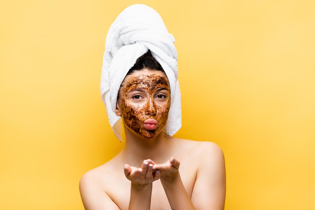 krásná žena s ručníkem na hlavě a kávová maska na tváři foukání polibek izolované na žluté - Fotografie, Obrázek