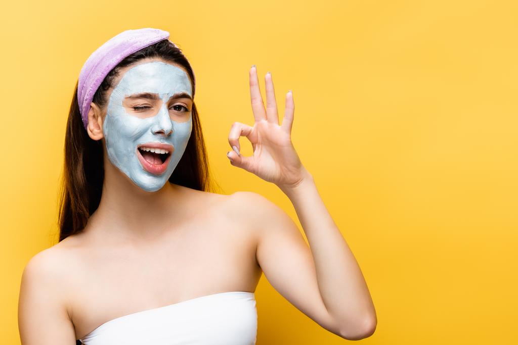 beautiful woman with clay mask on face showing okay isolated on yellow - Photo, Image