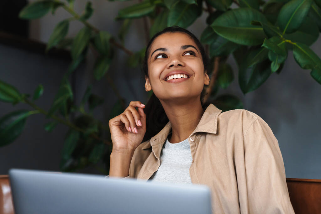 Sonriendo mujer afroamericana que trabaja con el ordenador portátil mientras está sentado en la cafetería - Foto, Imagen