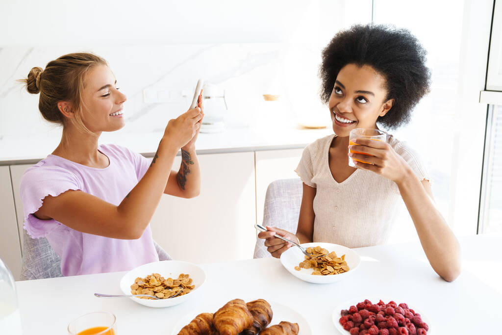 Pěkné veselé multikulturní dívky fotit na mobilu při snídani spolu doma kuchyně - Fotografie, Obrázek