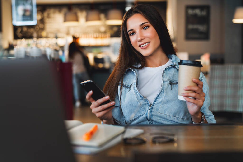 Primer plano. Hermosa joven mujer de negocios sonriente se sienta frente a la computadora portátil, sostiene la taza de café, utiliza el teléfono inteligente para el trabajo - Foto, Imagen
