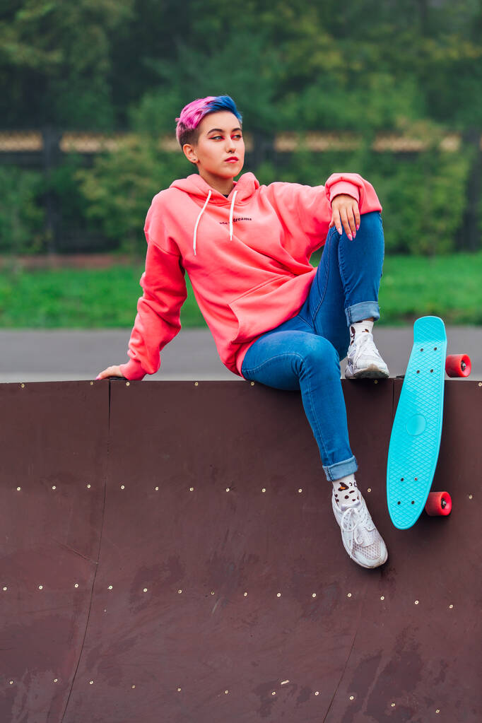 Portrét trendy hezká mladá dívka s krátkými barevnými vlasy a nos piercing oblečený v růžové mikiny a džíny sedí vedle skateboard soud s její modré plastové skateboard. - Fotografie, Obrázek