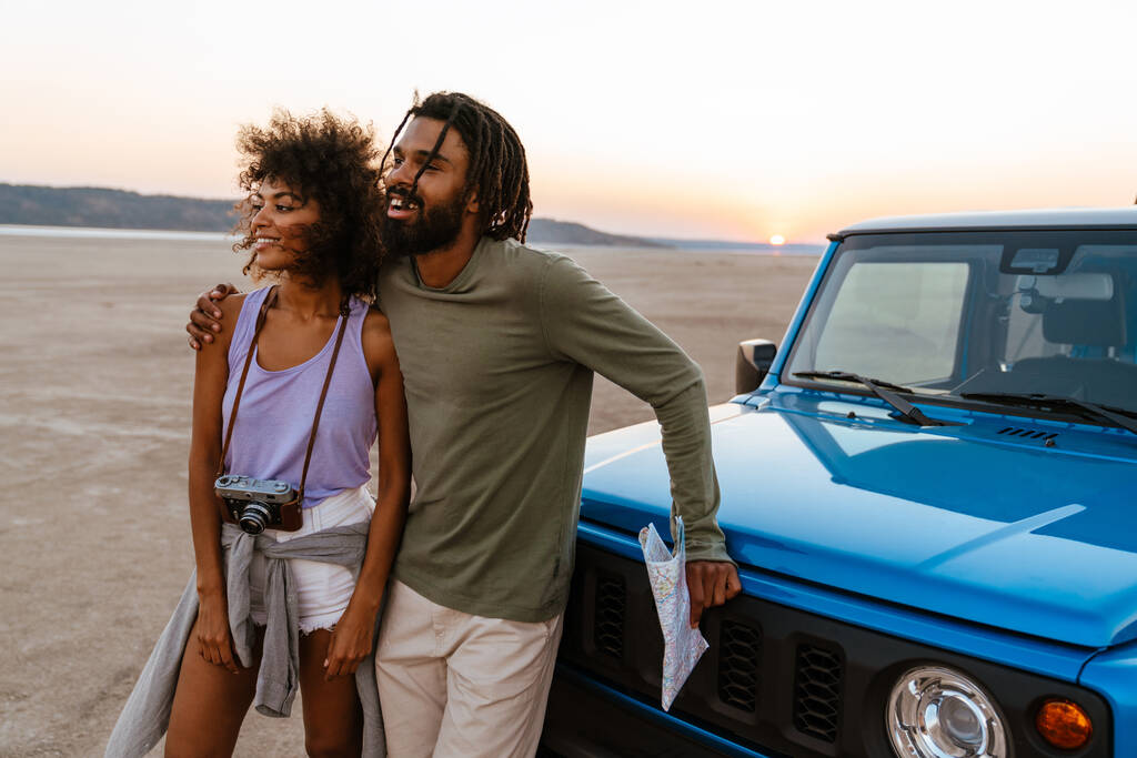 Радісна африканська подружня пара обіймає і посміхається під час подорожі з автомобілем у пустелі. - Фото, зображення