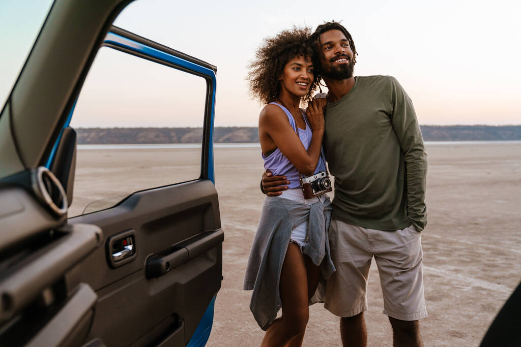 Изображение счастливой африканской пары, обнимающей и улыбающейся во время путешествия на машине по пустыне - Фото, изображение