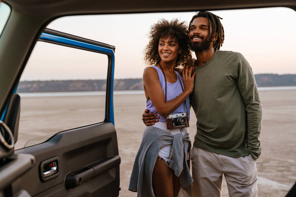 Imagen de la alegre pareja afroamericana abrazándose y sonriendo mientras viaja con el coche en el desierto - Foto, Imagen
